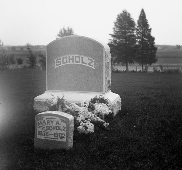 Scholz family monument located at Oak Hill Cemetery.  Flowers adorn the ground in front of the Monument.  A smaller tombstone labeled with the name Mary A. Scholz is stand in front of the larger family monument.