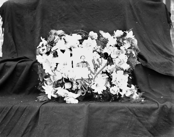 Funeral flower arrangement for Mrs. Henry Scholz. Her portrait is at the center of the arrangement with the word "Mother" written below it. The arrangement is propped up against a black backdrop.