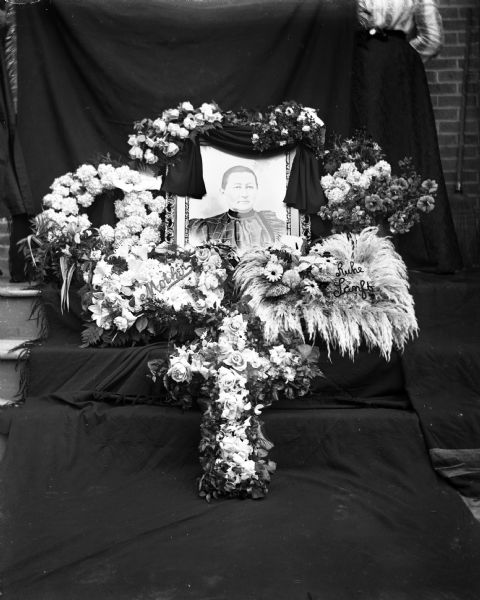 Portrait of Mr. Henry Scholz draped in a black cloth, surrounded by several funeral floral arrangements. The portrait is propped up against steps which are covered in a black backdrop. A woman in the background is helping to support the backdrop.