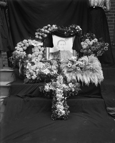 Portrait of Mrs. Henry Scholz draped in a black cloth, surrounded by floral arrangements for her funeral. The portrait and arrangements are propped up against several steps covered in a black backdrop. Two women are standing atop the stairs supporting the backdrop.