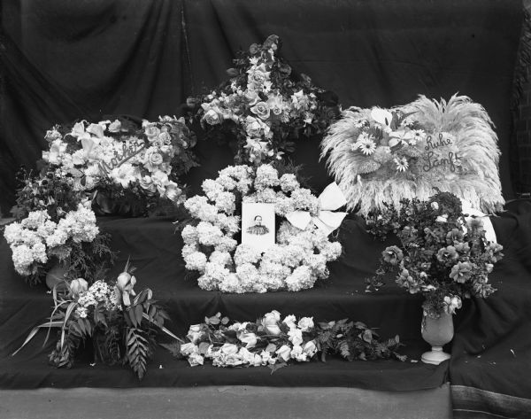 Eight floral arrangements for the funeral of Mrs. Henry Scholz arranged on a small staircase covered in a black backdrop. A small portrait of her has been placed in the center arrangement.