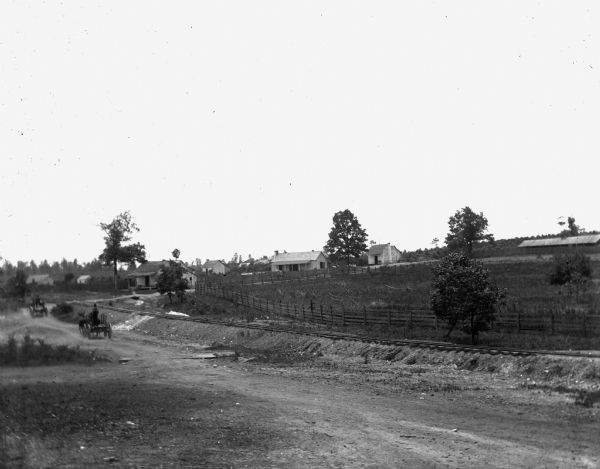 View of road along an African American settlement. A road is leading up to several houses on the left and a fenced-in field on the right. Two horse-drawn carts are traveling along the road. Railroad tracks are running parallel to the road on the right.
