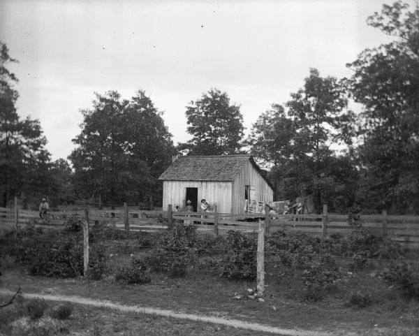 View over fence and garden towards an African American home and farm. The small home is standing along a tree line with the fenced-in garden stretching along the front of the house. Paul C. Goetsch is standing next to the house holding a baby. A women and small child are standing next to him, while another small child is sitting atop the garden fence.