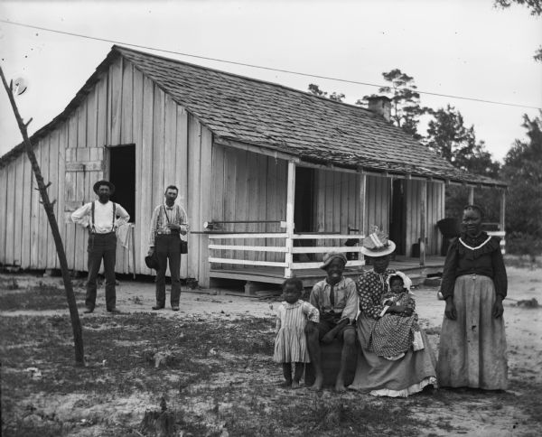 Alexander Krueger and E.S. Goetsch visiting a home in Shelby County, Alabama. The family, two women, two children, and a baby, are posing in front of the house, while the two men are standing closer to the home behind them on the left.