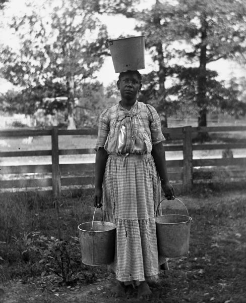Barefoot woman standing in front of a fence carrying three water buckets. She is holding a bucket in each hand and balancing another on her head.