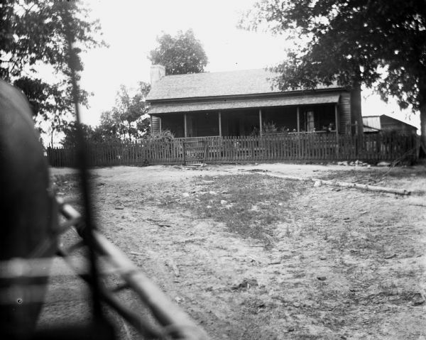 View from a horse-drawn vehicle of an old home that was built prior to the Civil War. A picket fence surrounds the house. There is a full-length porch on the front of the house, and behind the house on the right are sheds and outbuildings.