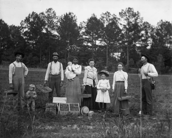 Outdoor group portrait of chidren and adults, standing in the strawberry field of Paul C. Goetsch after picking strawberries. Four people are holding crates of freshly picked strawberries in their hands, and large crates are sitting on the ground in front of them. From left to right are: Paul C. Goetsch, Russel Goetsch, Ernst Goetsch, unidentified woman holding Dorothy Goetsch, Mrs. Ernst Goetsch, Earnie Goetsch, Mrs. Paul C. Goetsch, and Alexander Krueger. Alexander is operating the camera by pulling a string he is holding in his hand.