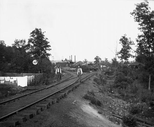 Alexander Krueger and Ernst Goetsch standing along a railroad track leading to a sawmill. The mill stais at the end of the tracks, which is lined with trees. Two women are standing on the tracks in the far background. Laundry is drying on a fence on the left.