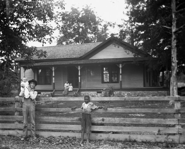 Paul C. Goetsch and family posing in front of their home. Mrs. Paul C. Goetsch and Russel Goetsch are sitting on the steps of the front porch. Paul is holding Dorothy and is standing along the fence in front of the house. Also standing against the fence is a boy named Hosley, who picks strawberries for the Goetsches.