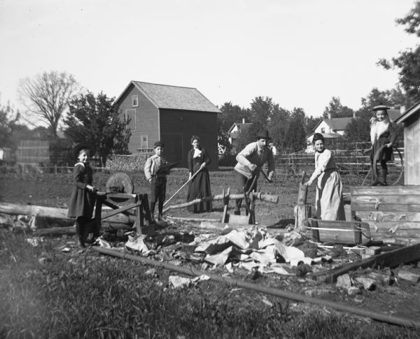 Henry Miller with his wife, three children, and sister-in-law, Lena Jaeger, chopping wood. Henry is sawing a log, while a woman is cutting wood with an axe next to him. Another woman is using a hoe behind him. On the far right, a young girl is standing on an elevated platform with a hoop, while on the far left her brother is holding a board with sticks piled on top. A girl is standing next to him on the left sharpening a blade on a sharpening stone. The ground in front of them is littered with fresh cut wood. Several houses and outbuildings are in the background.