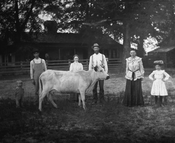 Goetsch family standing with a cow in front of the home of Paul C. Goetsch. From left to right are: Russel Goetsch, Paul C. Goetsch, Mrs. Paul C. Goetsch, Ernst Goetsch, Mrs. Ernst Goetsch, and Earnie Goetsch. A woman is standing on the porch of the house in the background.