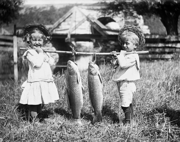 Jennie and Edgar Krueger holding a pole between them on their shoulders, with two carp strung up on it. They are standing on a bank next to a river, with a bridge in the background.