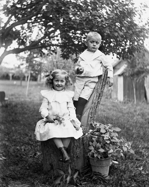 Outdoor portrait of Jennie and Edgar Krueger, posing on a log cut into a chair. Jennie is sitting on the log seat holding a bouquet of flowers, while Edgar is standing behind her leaning against the back of the chair holding an apple. A potted plant is sitting on the ground next to the log.