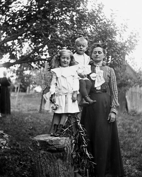 Outdoor family portrait of Jennie, Edgar, and Florentina Krueger posing with a log that has been carved into a chair. Jennie is standing on the seat of the chair, leaning against the back holding a bouquet of flowers, while her mother, Florentina, is standing next to the chair. Edgar is sitting between them atop the back of the chair holding an apple. A plant with leaves is in front of the chair. Mary Krueger is standing in the far background on the left, next to a tree.