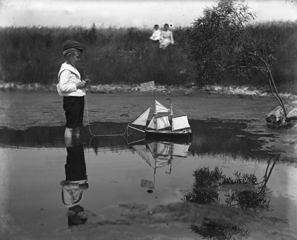 Edgar Krueger standing in a lime kiln pond playing with a toy sailboat. An American flag is on top the mast of the sailboat. Florentina and Jennie Krueger are sitting on a hill along the shoreline in the background.