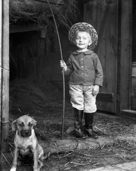 Edgar Krueger standing in the doorway of a hay barn holding a fishing pole in his right hand. He is wearing a straw hat and rubber boots. A puppy is sitting in the doorway near him on the left.