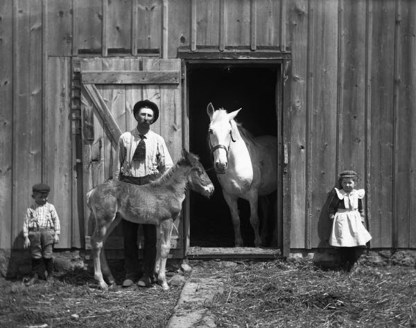 Edgar, Alexander, and Jennie Krueger standing outside a barn. A young colt named "Frank" is standing in front of Alexander. The colt's mother, "Flora," is standing looking out of the open barn door.