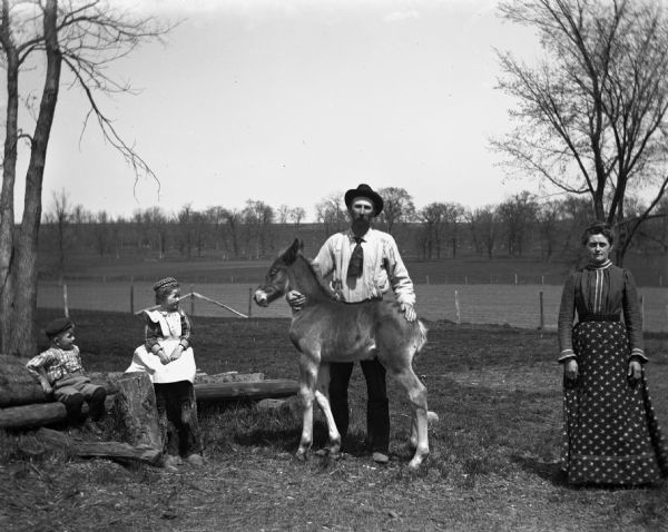 Alexander Krueger family posing in a pasture with a colt named Frank. Alexander Krueger is standing behind the colt with one hand under its neck and the other on its hindquarter. Jennie and Edgar Krueger are sitting on a pile of timber on the left, while Florentina is standing on the right off to the side.