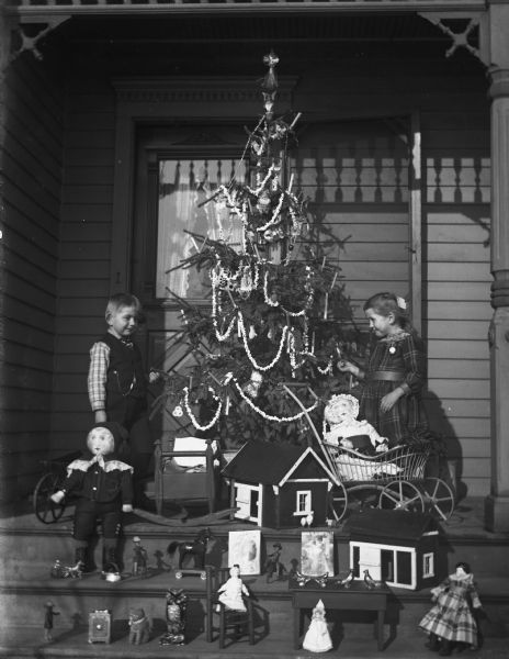 Jennie and Edgar Krueger standing on the porch with a decorated Christmas tree between them. Toys they received as presents are displayed on the steps in front of them.