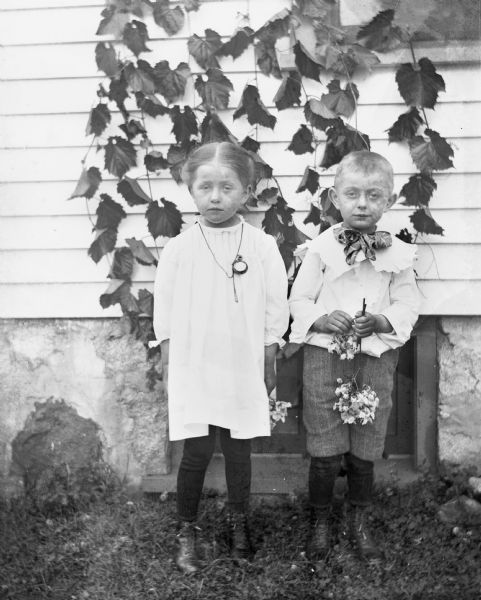 Outdoor portrait of Jennie and Edgar Krueger standing in front of the side of a house. Jennie is wearing a pocket watch in her front pocket with the chain worn around her neck. Both children are holding flowers in their hands. Ivy drapes the side of the house.