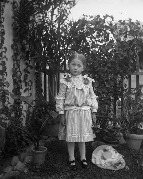Outdoor portrait of Jennie Krueger standing along a garden fence next to a building holding flowers in her left hand. Her hat, decorated with ribbons and flowers, is placed on the ground beside her. Potted plants are lining the border of the yard.