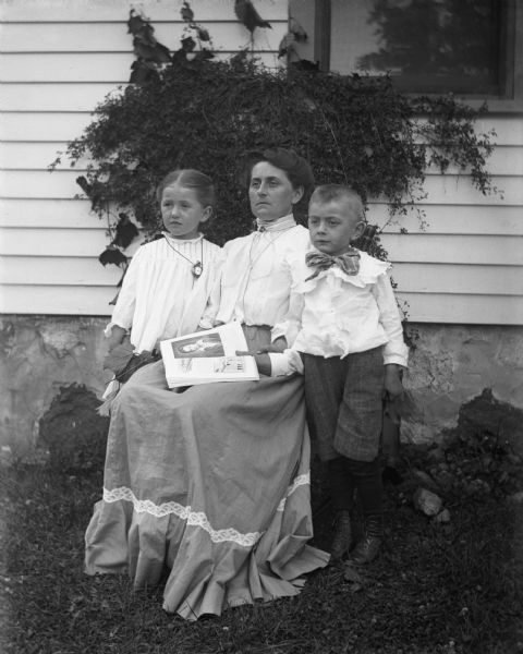 Outdoor portrait of Florentina Krueger sitting in a chair with her two children, Jennie and Edgar Krueger, standing on either side of her. A book is lying open in her lap.