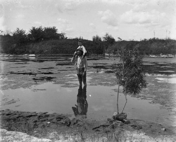 Alexander Krueger standing in the middle of a quarry pond with his son, Edgar Krueger, sitting on his shoulders.