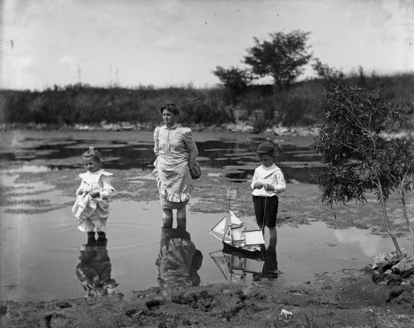 Jennie, Florentina, and Edgar Krueger standing in a quarry pond. Jennie and Florentina are holding up their dresses, while Edgar is playing with a toy sailboat.