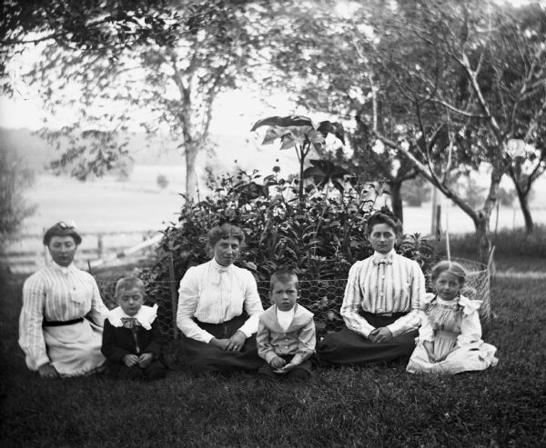 Outdoor group portrait of three women and three children sitting in a yard in front of a small fenced in flower bed. From left to right: Sarah Krueger, Edgar Krueger, Lydia Lally, Edwin Lally, Florentina Krueger, and Jenni Krueger.