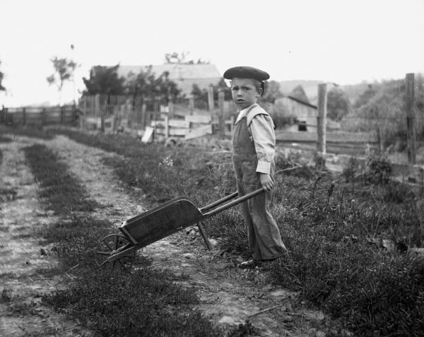 Edwin Lally standing on a dirt road next to a fence pushing a wheelbarrow. Farm buildings and haystacks are beyond the fence.