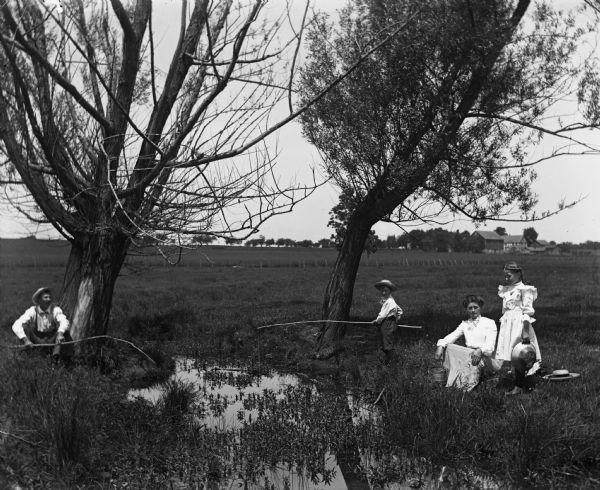 Alexander Krueger and family fishing in a small pond on their farm. Alexander and Edgar are using sticks as fishing poles, while Florentina and Jennie are sitting and standing along the edge holding a pail and a pan. In the far background are farm buildings.