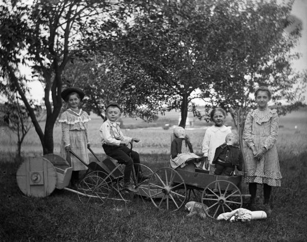 Esther Bigalk Clark, Edgar Krueger (on tricycle), Jennie Krueger, and Josie Bigalk Wendorf playing with wagons, dolls and a toy wheelbarrow outdoors.