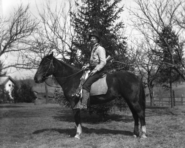Alexander Krueger riding a horse named Jim, while holding a rifle. Alexander is wearing a hat, a bandana around his neck, striped shirt, shotgun shell belt, and laced-up gaiters over his boots. A dead rabbit is strung-up along the front of the saddle by its back feet, and is draping down the side of the horse.