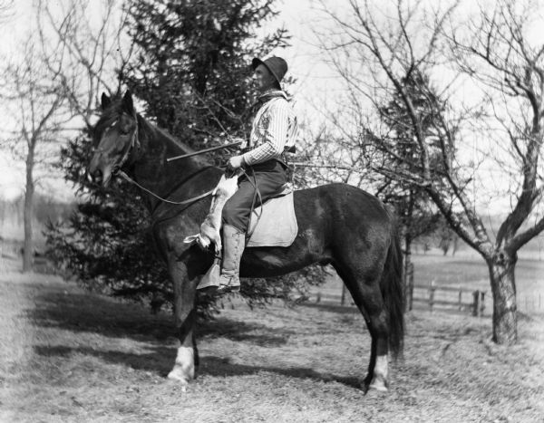 Alexander Krueger riding a horse named Jim, while holding a rifle. Alexander is wearing a hat, bandana around his neck, striped shirt, shotgun shell belt, and laced-up gaiters over his boots. A dead rabbit is strung-up along the front of the saddle by its back feet, and is draping down the side of the horse.