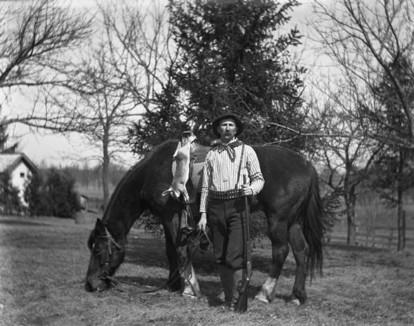 Alexander Krueger standing in front of his horse, Jim, while holding a rifle. Alexander is wearing a hat, a bandana around his neck, striped shirt, shotgun shell belt, and laced-up gaiters over his boots. A dead rabbit is strung-up along the front of the saddle by its back feet, and is draping down the side of the horse.