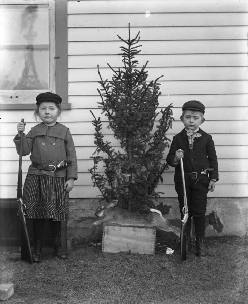 Jennie and Alexander Krueger standing against the side of a house holding up rifles. Each of them is wearing a belt with a pistol propped inside. A dead rabbit is laid out atop a box in front of a small pine tree that is standing between them, and two dead birds are hanging from one of the branches.