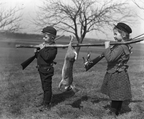 Jennie and Edgar Krueger suspending a tree limb on their shoulders on which is hanging a dead rabbit and two birds. Each of them is holding a rifle over their right shoulder, and they have pistols stuck in their belts.