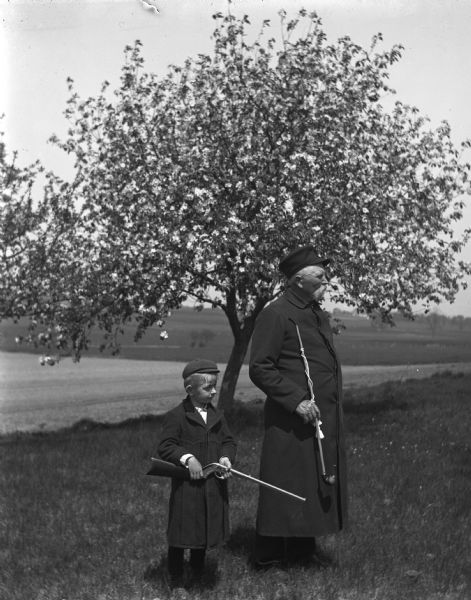 Edgar and August Krueger standing in front of a blossoming apple tree. Edgar is holding a rifle, while August is holding a long pipe with a cord and tassel. Both are dressed in long coats.