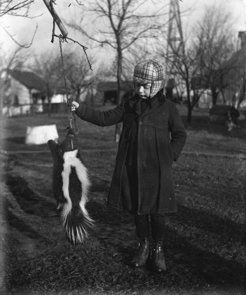 Edgar Krueger holding onto an animal trap, hanging from a tree branch, with a dead skunk trapped by a front paw. Farm buildings are in the background.