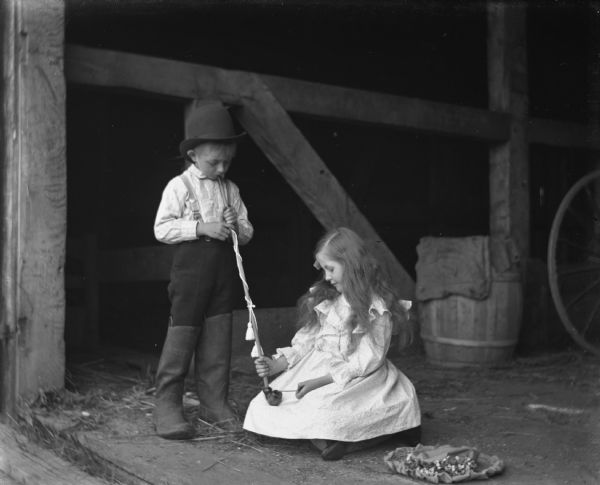 Jennie and Edgar Krueger playing with August's long pipe decorated with a cord and tassel in a barn. Edgar is holding the pipe up to his mouth while Jennie is lighting it. Edgar is dressed in Pomeranian attire.