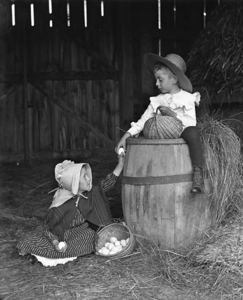 Jennie and Edgar Krueger sitting in a barn, each with a handmade basket filled with eggs. Edgar is sitting atop a barrel with his right hand on an egg, while his sister is sitting on the ground below reaching up with her left hand to hold the bottom of the egg. Jennie is dressed in Pomeranian attire.