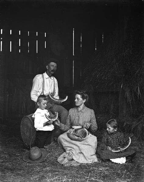 Alexander Krueger family eating watermelon in a hay barn. Alexander, Edgar, and Jennie are each holding a slice of watermelon, while Florentina is cutting up slices of the melon for them.