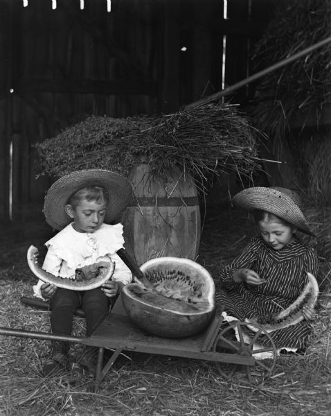 Edgar and Jennie Krueger eating watermelon in the hay barn. The watermelon and a knife are sitting between them on a small wheelbarrow.