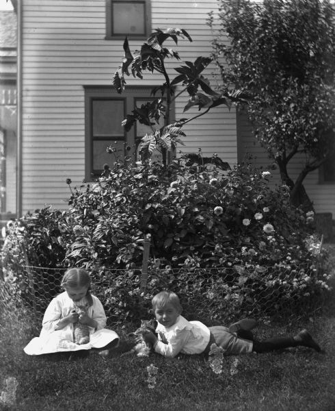 Jennie and Edgar Krueger playing with kittens in the yard next to a fenced-in flower bed near the side of a house. Each of them is holding a kitten.