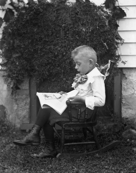 Outdoor portrait of Edgar Krueger sitting in a rocking chair reading a book next to a house.  He has the book he is reading open in his lap. Ivy is growing on the side of the house as part of the backdrop.