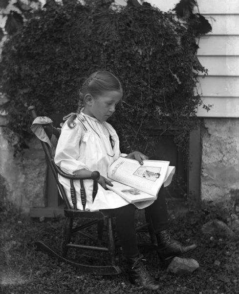 Outdoor portrait of Jennie Krueger sitting in a rocking chair, next to a house, reading a book.  The book she is looking at is open and resting in her lap.  Ivy is growing along the side of the house as part of the backdrop.