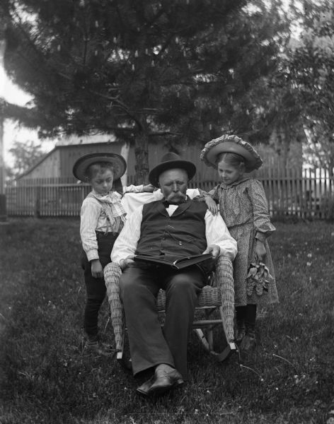 Outdoor portrait of August, Jennie, and Edgar Krueger in the yard. August is sitting in a wicker rocking chair with a book open in his lap, while the twins are standing behind him looking over his shoulder.  Jennie is holding lilacs in her left hand.