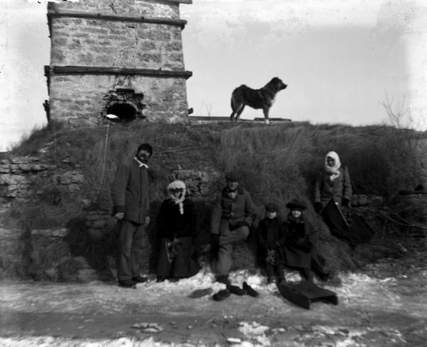 Members of the Krueger and Buelke family standing below an old kiln during winter. Some of them are holding ice skates and a sled is in front of the twins. A dog is standing on top of the hill in the background. From the left are: Alexander Krueger, Sarah Krueger, Ernie Buelke, Edgar and Jennie Kruger, and Rose Buelke.