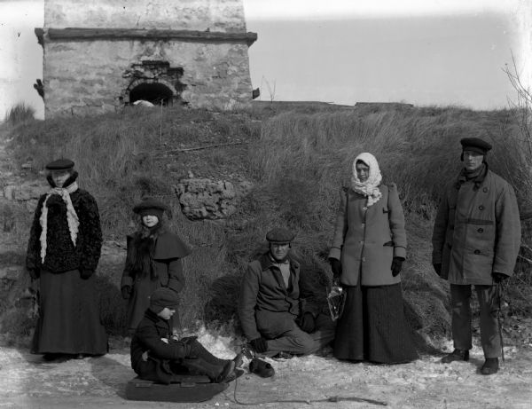 Group posing next to the quarry pond with the old kiln on the hill above them.  Several people in the group are holding ice skates. From the left are: Sarah Krueger, Jennie Krueger, Ernie Buelke, Rose Buelke, and William Buelke.  Edgar Krueger is sitting in front of them on a sled.