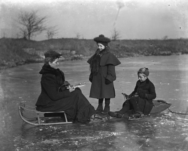 Florentina, Jennie, and Edgar Krueger preparing for ice skating on the quarry pond. Florentina and Edgar are sitting on sleds on the ice while attaching blades to their shoes. Jennie is standing between them.